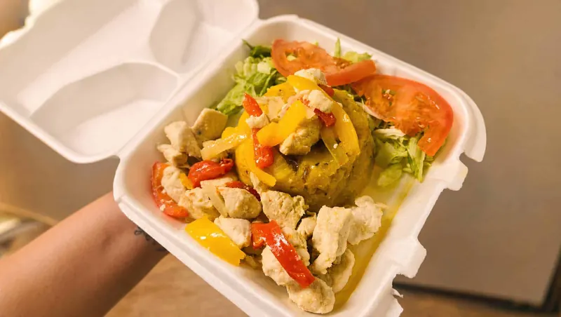 Mofongo topped with seasoned pollo, sweet peppers, lettuce and tomato in a to-go container